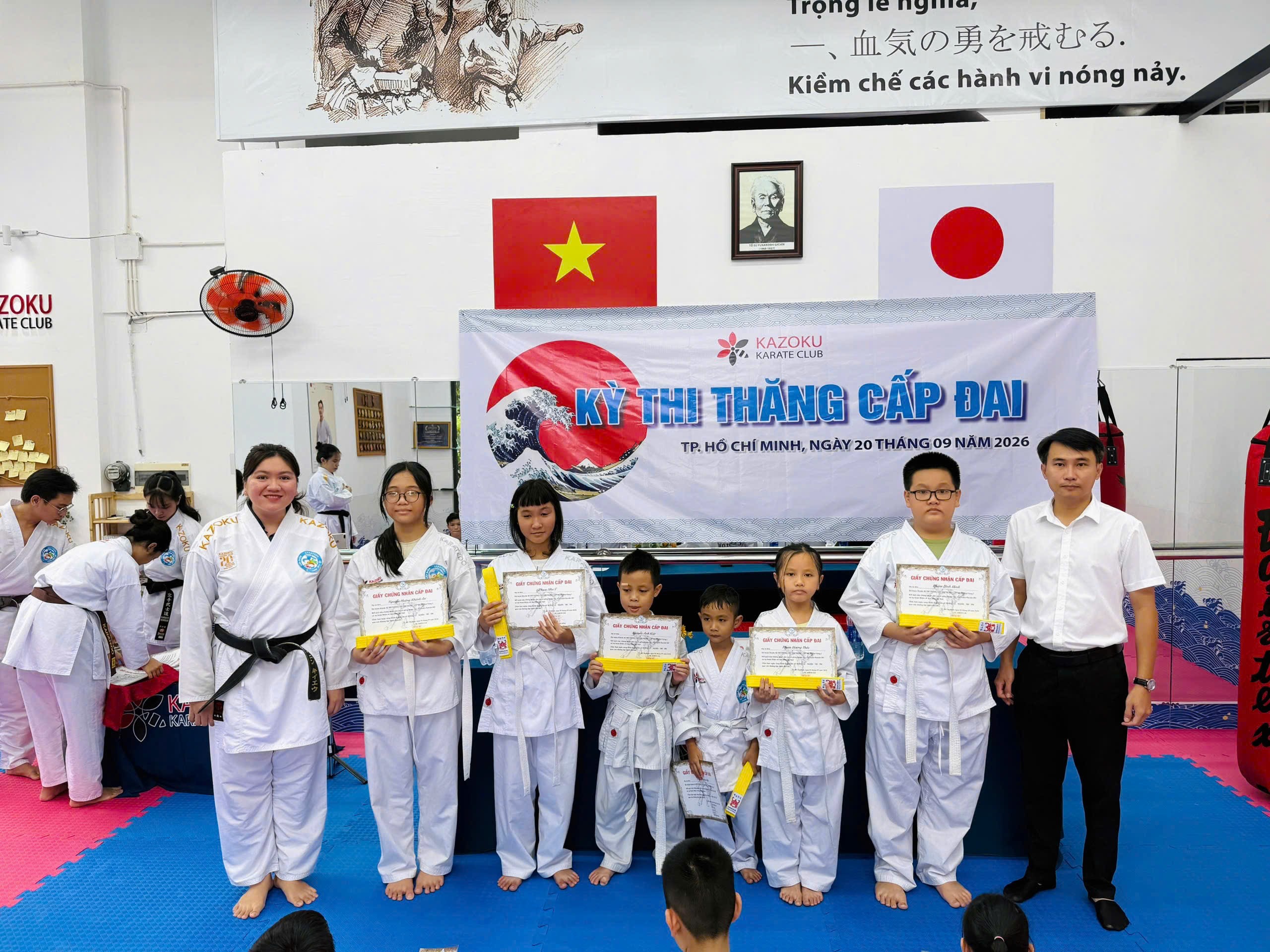 Một nhóm võ sinh mặc võ phục đứng trước phông Kỳ thi thăng cấp đai; một số người cầm giấy chứng nhận và đai vàng, bên cạnh có một người mặc áo sơ mi trắng.