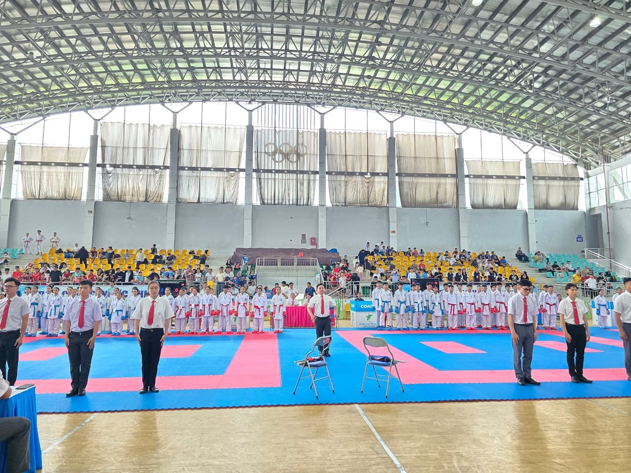 Các vận động viên karate mặc võ phục trắng xếp hàng trên thảm đấu đỏ và xanh trong nhà thi đấu, phía trước là những người mặc áo sơ mi trắng và cà vạt đỏ.
