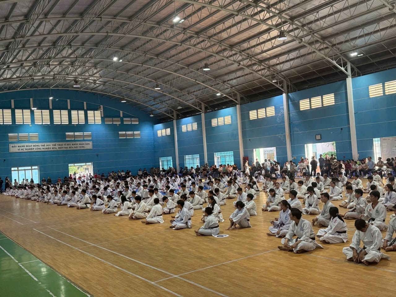 Đông võ sinh mặc võ phục trắng ngồi thành nhiều hàng trên sàn gỗ của nhà thi đấu trong nhà, phía sau có người đứng dọc theo tường.