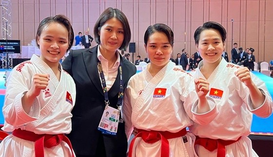 Huấn luyện viên Nguyễn Hoàng Ngân mặc trang phục tối màu đứng cùng ba vận động viên Karate nữ Việt Nam mặc võ phục trắng và đeo đai đỏ tại một địa điểm thi đấu.