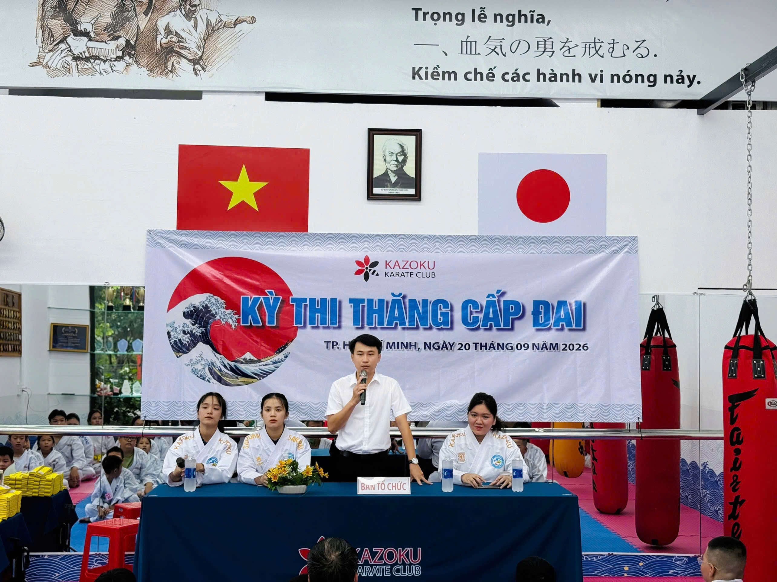 Một người cầm micro đứng sau bàn có biển Ban tổ chức, phía trước phông Kỳ thi thăng cấp đai ghi ngày 20 tháng 09 năm 2026; các võ sinh mặc võ phục ngồi phía sau.
