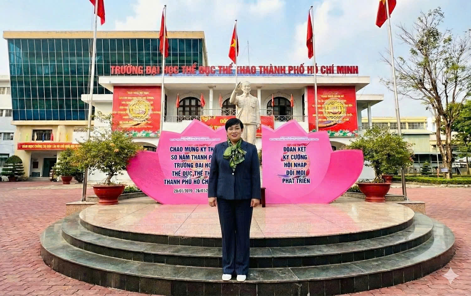 Bà Huỳnh Thị Ngọc Phượng đứng trước Trường Đại học Thể dục Thể thao Thành phố Hồ Chí Minh, phía sau là cờ và pa-nô kỷ niệm.