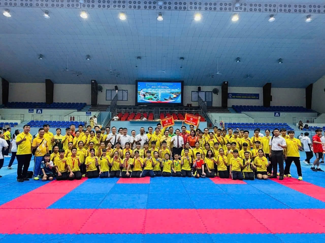 Nhóm đông người mặc áo vàng chụp ảnh tập thể trên thảm đấu đỏ và xanh trong nhà thi đấu; một số người ở hàng sau cầm cờ đỏ.
