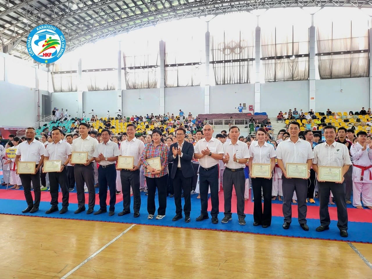 Một dãy người ngồi sau bàn phủ khăn đỏ, phía trước phông nền của giải karate năm 2026 trong nhà thi đấu.