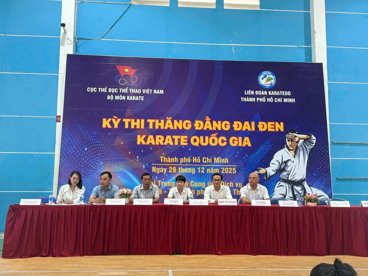 Sáu người ngồi sau bàn phủ khăn đỏ trước phông nền của Kỳ thi thăng đẳng đai đen Karate quốc gia, ghi ngày 28/12/2025 và tên Liên đoàn Karatedo TP.HCM.